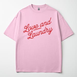 Puede incluir: Camiseta rosa claro con la frase "Love and Laundry" en rojo, escrita en cursiva. La camiseta tiene mangas cortas y cuello redondo. La tela parece suave y cómoda.