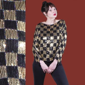 Puede incluir: Una blusa negra y dorada con lentejuelas y un patrón de cuadros. La blusa tiene mangas largas y un corte holgado.