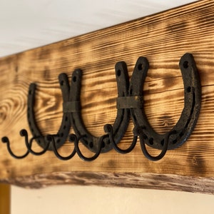 May include: Three black metal horseshoes mounted on a rustic wooden beam, each horseshoe has two hooks for hanging items.