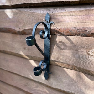 Könnte beinhalten: Schwarzer Wandleuchter aus Schmiedeeisen mit zwei Kerzenhaltern und dekorativen Schnörkeln. Der Leuchter ist an einer Holzwand befestigt. Der Leuchter ist etwa 30 cm hoch.