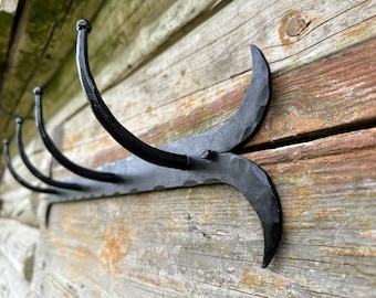 May include: Black metal coat rack with four hooks. The hooks are curved and have rounded ends. The rack is mounted on a wooden wall.