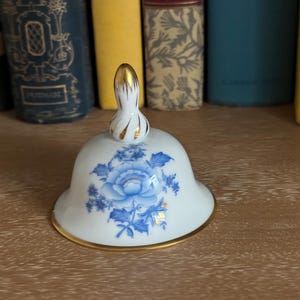 May include: A white porcelain bell-shaped lid with a gold-trimmed edge and a gold finial. The lid features a blue floral design. The bell is resting on a wooden surface, with a backdrop of colorful books.