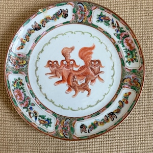 May include: A round, antique porcelain plate with a red rim and a white center. The center features a group of orange-red mythical creatures. The border is decorated with colorful floral and butterfly motifs, with green, pink, and yellow accents.
