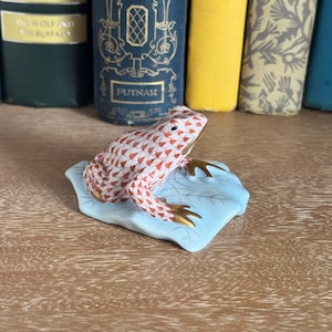 May include: A ceramic frog figurine with a red and white patterned body and gold-colored feet, resting on a light blue leaf. The frog is a decorative item, likely for display.