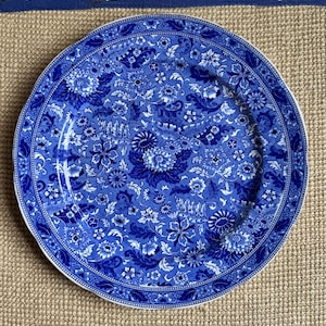 May include: A round ceramic plate featuring a dense, intricate blue and white floral pattern. The design covers the entire surface with various stylized blossoms, leaves, and scrolling vines, framed by a decorative border along the outer edge.