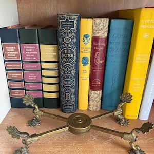May include: A collection of vintage books with colorful spines, including titles like "The Sketch Book" and "Gone With The Wind," arranged on a wooden shelf. A brass book stand with decorative details is in the foreground.