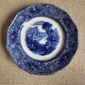 May include: A blue and white decorative plate with a landscape scene in the center. The plate has a scalloped edge and floral detailing. The plate is made of ceramic and is likely vintage.