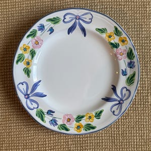 May include: White ceramic plate with a blue rim and floral design. The plate features a pattern of blue bows, pink and yellow flowers, and green leaves. A decorative piece for serving food.