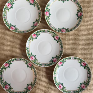 May include: Five white porcelain saucers with a gold rim and a floral design. Each saucer features a border of pink roses and green leaves. The saucers are arranged on a textured, neutral-toned surface.