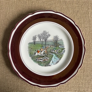 May include: A round ceramic plate with a scalloped edge featuring a wide, deep maroon border. The center of the plate displays a detailed, colorful illustration of a fox hunting scene with riders on horseback, hounds, and a stream in a landscape.