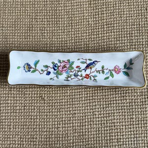 May include: A rectangular white ceramic dish with a gold trim. The dish is decorated with a floral and bird design in shades of blue, pink, and green. The dish is approximately 8 inches long.