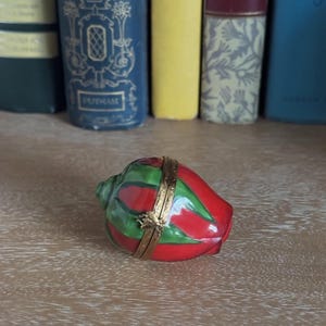 May include: A shell-shaped trinket box, painted with red and green stripes, featuring a gold-coloured metal hinge and clasp. The box sits on a wooden surface, with a backdrop of vintage books. The box is approximately 7 cm in length.