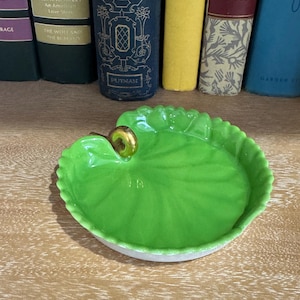 May include: A bright green, leaf-shaped ceramic dish with a gold accent. The dish has a white base and is set against a backdrop of books. The dish is likely used for holding small items.