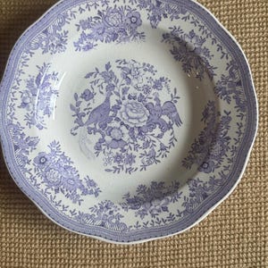 May include: A round ceramic plate with a scalloped edge featuring a white base and a purple transferware pattern. The design includes central floral arrangements, two birds, and a decorative border of repeating flowers and geometric lines.
