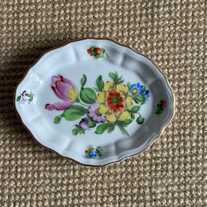 May include: A small, white, oval-shaped dish with a scalloped edge and gold trim. The dish is decorated with a colorful floral design featuring pink, yellow, and blue flowers with green leaves. A decorative piece for the home.