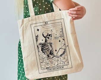 Puede incluir: Una bolsa de lona blanca con una ilustración en blanco y negro de un esqueleto leyendo un libro. La ilustración está rodeada de flores y estrellas. El texto "XI THE READER" está impreso debajo de la ilustración.