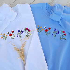 May include: Two sweatshirts with embroidered flowers and a bee. One sweatshirt is white and the other is light blue.