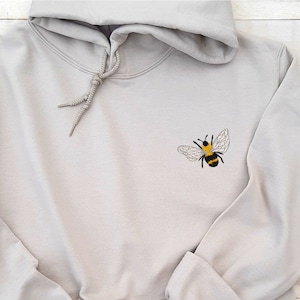 May include: A light grey hoodie with a small embroidered bee on the left chest. The bee is black and yellow with transparent wings.