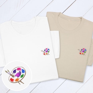 以下が含まれることがあります： 白とベージュの2枚のTシャツ。それぞれに、カラフルな絵の具の飛沫と筆が描かれた、小さな刺繍のアーティストパレットのデザインが施されています。パレットのデザインには、赤、オレンジ、黄色、緑、紫、青が含まれています。