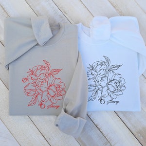 May include: Two sweatshirts with a floral design. One sweatshirt is gray with a red floral design and the word "Peony" printed below. The other sweatshirt is white with a black floral design and the word "Peony" printed below.
