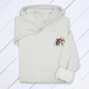 May include: A light gray hoodie with a drawstring hood and white cuffs. The hoodie features a small embroidered design of a gray mouse holding red berries on the upper left sleeve. The hoodie is made of a soft, comfortable material.