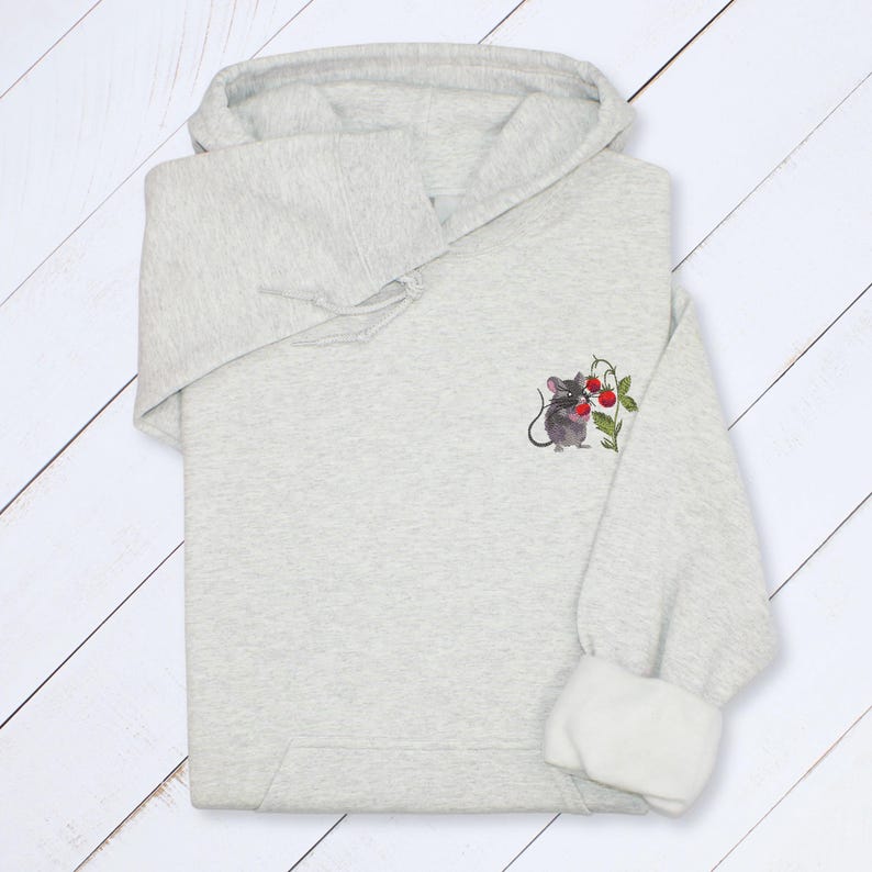 May include: A light gray hoodie with a drawstring hood and white cuffs. The hoodie features a small embroidered design of a gray mouse holding red berries on the upper left sleeve. The hoodie is made of a soft, comfortable material.