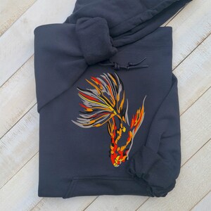 Peut inclure: Un sweat-shirt bleu fonc&eacute; avec un motif de poisson brod&eacute; color&eacute; sur le devant. Le poisson est stylis&eacute; et a une longue queue avec des accents orange, jaune et gris.