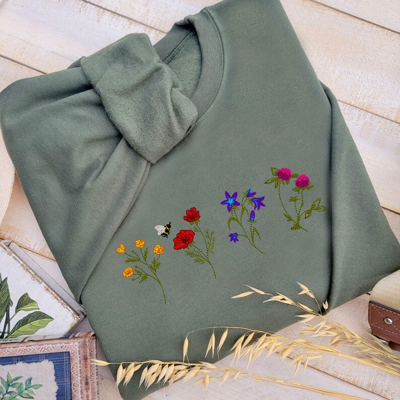 May include: A green sweatshirt with embroidered flowers and a bee. The flowers are red, blue, purple, and yellow.
