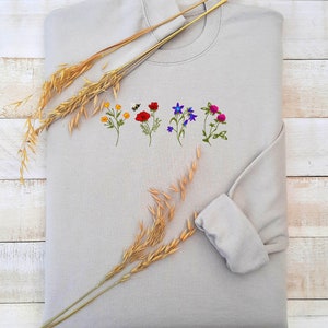 May include: A light gray sweatshirt with embroidered flowers in shades of red, blue, yellow, and green. The flowers are arranged in a row across the center of the sweatshirt.