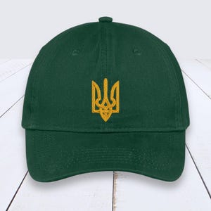May include: A dark green baseball cap with a gold embroidered emblem. The cap has a curved brim and a fabric strap closure. The emblem is a stylized trident, a symbol of Ukraine.