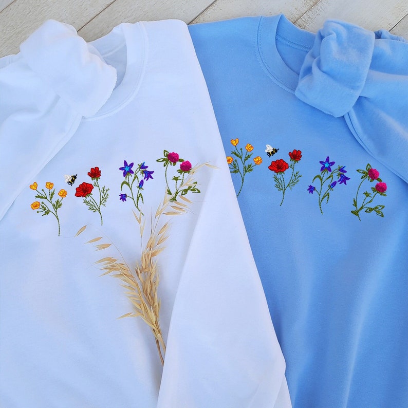 May include: Two sweatshirts with embroidered flowers and a bee. One sweatshirt is white and the other is light blue.