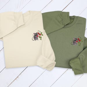 May include: Two long-sleeved sweatshirts, one cream and one olive green, are displayed. Each features an embroidered design of a gray mouse holding red berries. The sweatshirts are folded, showcasing the embroidered detail on the chest.
