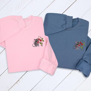 May include: Two long-sleeved shirts, one pink and one blue, each featuring an embroidered design of a gray mouse holding red cherries. The shirts have a crew neck and cuffed sleeves, perfect for casual wear.