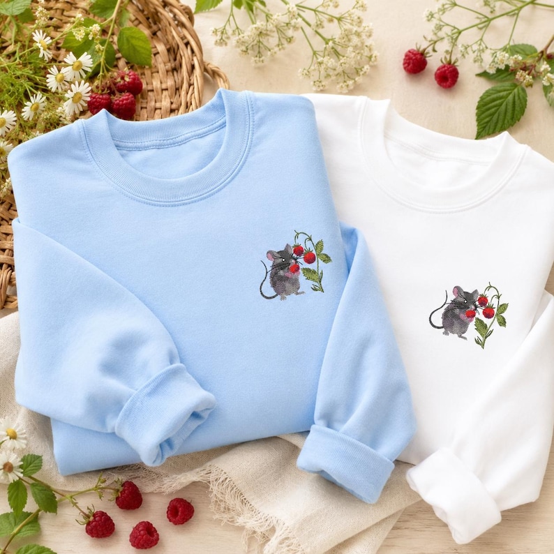 May include: Two sweatshirts, one light blue and one white, each featuring an embroidered design of a gray mouse holding red berries. The sweatshirts have long sleeves and a crew neck. The background includes flowers and raspberries.