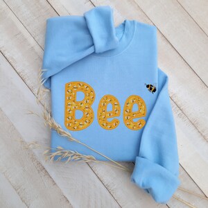 May include: A light blue sweatshirt with the word "Bee" spelled out in yellow letters. Each letter is made up of a pattern of small bees. A single bee is flying above the word.