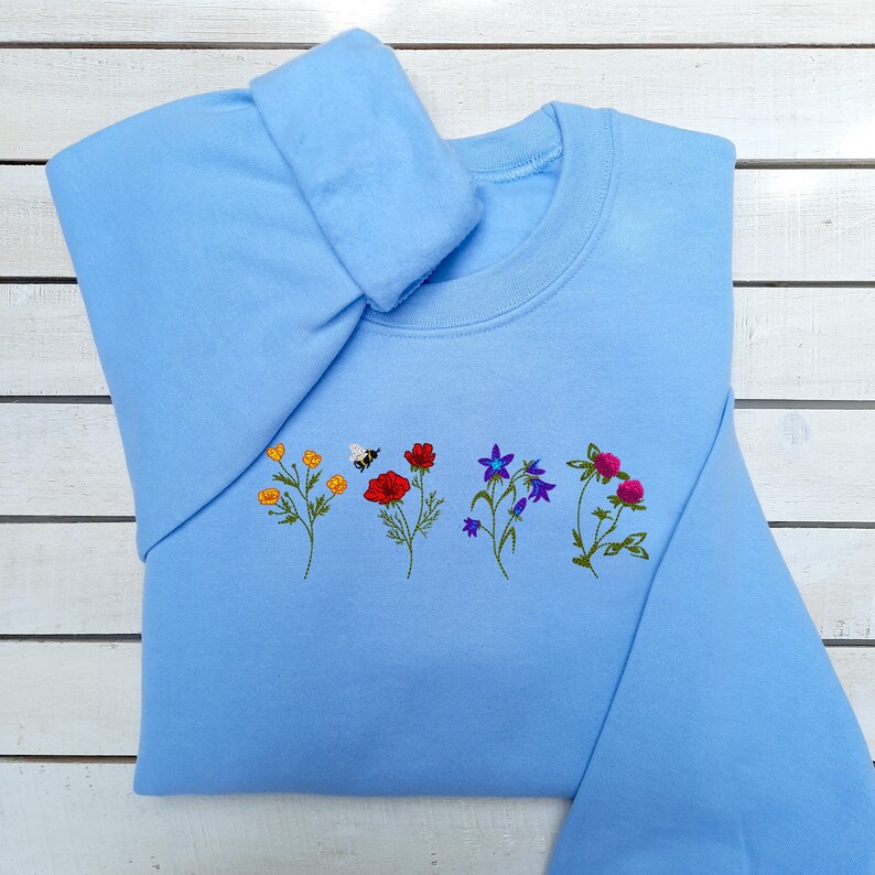 May include: A light blue sweatshirt with embroidered flowers and a bee on the front.