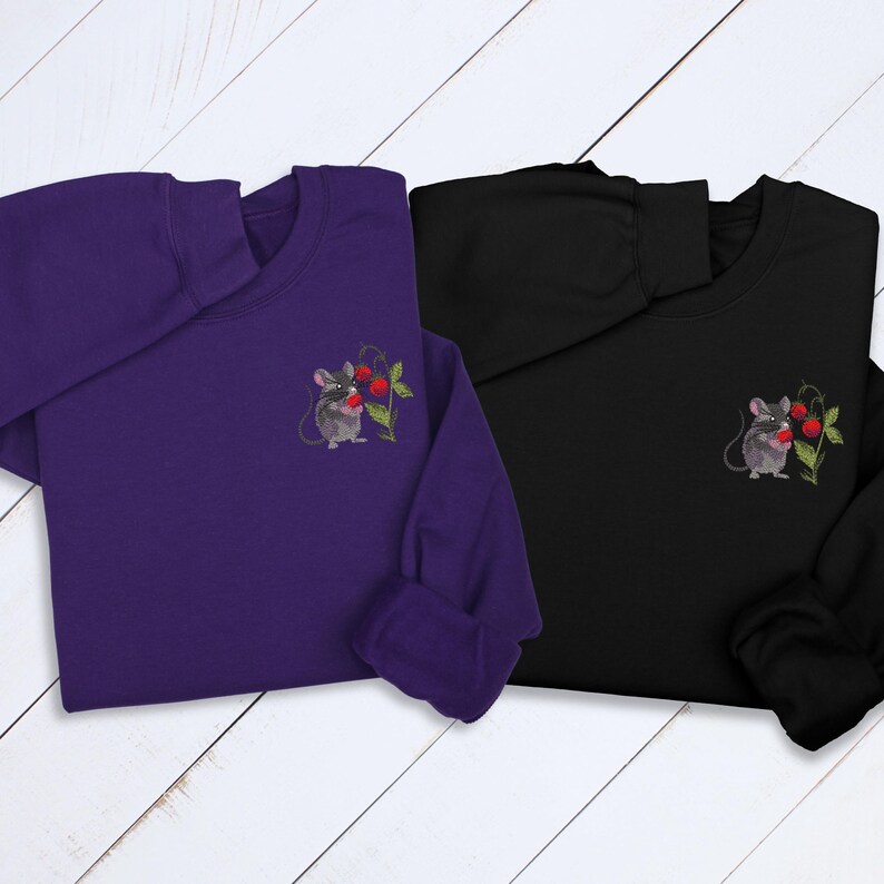 May include: Two sweatshirts, one purple and one black, are displayed. Each has a small embroidered design of a mouse holding a red berry sprig on the chest. The sweatshirts are long-sleeved and have a crew neck.
