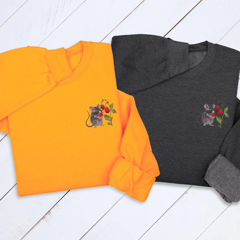 May include: Two sweatshirts, one orange and one charcoal gray, each featuring an embroidered design of a mouse holding cherries. The mouse and cherry design is on the left chest area of each sweatshirt. The sweatshirts have long sleeves and a crew neck.