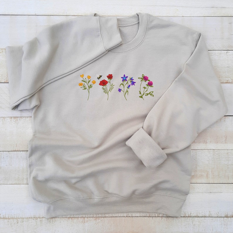 May include: A light gray sweatshirt with a row of embroidered flowers in various colors, including red, yellow, blue, and purple.
