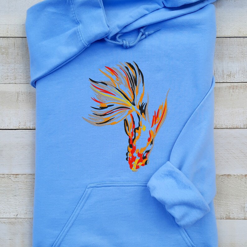 Peut inclure: Un sweat-shirt bleu clair avec une image brod&eacute;e d'un poisson rouge avec des d&eacute;tails orange, rouge, jaune et noir.