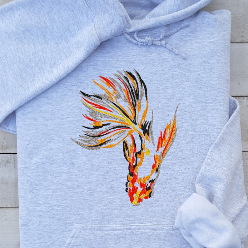 Peut inclure: Un sweat-shirt gris avec une image brod&eacute;e d'un poisson rouge avec des d&eacute;tails orange, rouge et noir.