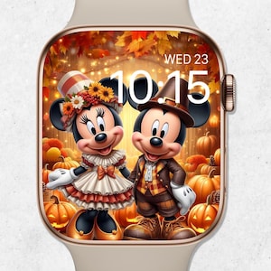 May include: A beige smartwatch with a screen displaying Mickey and Minnie Mouse in autumn attire, surrounded by pumpkins and fall foliage. The time is 10:15 AM, Wednesday, October 23rd. The watch has a gold-colored bezel and a beige band.