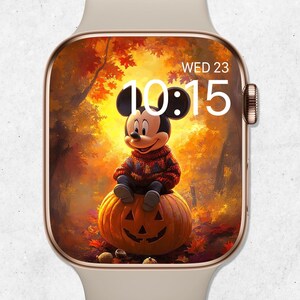 Pode incluir: Um smartwatch bege com moldura dourada exibe uma imagem com tema de outono. A tela mostra Mickey Mouse sentado em uma abóbora esculpida em um cenário de floresta. A hora é 10:15 e o dia é quarta-feira.