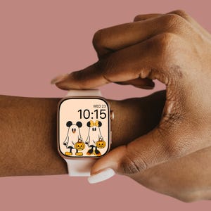 May include: A white smartwatch with a gold band displays a Halloween-themed watch face with Mickey Mouse and Minnie Mouse dressed as ghosts holding jack-o'-lanterns. The watch face also displays the time, 10:15, and the day, WED 23.