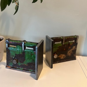 May include: A pair of L-shaped bookends constructed from industrial steel beams. Each piece features a weathered, rusted texture combined with patches of vibrant green paint and a smooth, polished metal vertical plate on the outer edge.