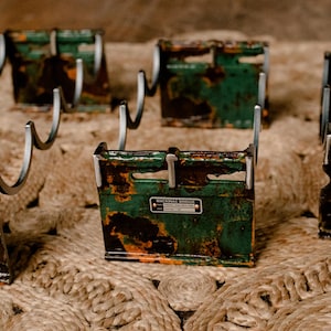 May include: Four vintage metal holders with hooks, painted in a green, black, and orange camouflage pattern. Each holder has a small metal tag with the text "GENERAL ELECTRIC" printed on it.
