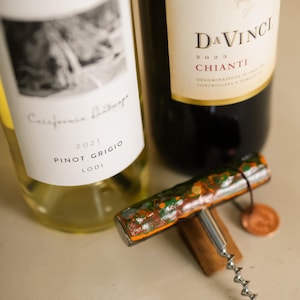 May include: Two wine bottles, one labeled "California Landscape Pinot Grigio Lodi 2021" and the other "DaVinci Chianti 2023." A colorful corkscrew with orange, green, and brown accents rests on a wooden base, with a small copper tag.