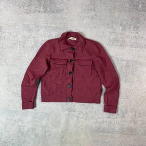 May include: A burgundy-colored jacket with a collared neckline and two front pockets. The jacket has long sleeves with folded cuffs and is fastened with dark buttons. The garment is made of a soft, textured material.