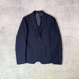 Puede incluir: Un blazer azul marino oscuro con solapa de muesca, dos botones frontales y dos bolsillos inferiores con solapa. La prenda incluye un bolsillo en el pecho, parches visibles en los codos de las mangas y un forro interior oscuro en el cuello.