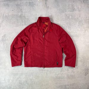 Puede incluir: Una chaqueta roja brillante y ligera con cierre de cremallera completo y cuello alzado. La chaqueta tiene mangas largas y un pequeño bolsillo en el lado inferior izquierdo. El interior es de color naranja en contraste. La chaqueta es de material liso, posiblemente nailon.